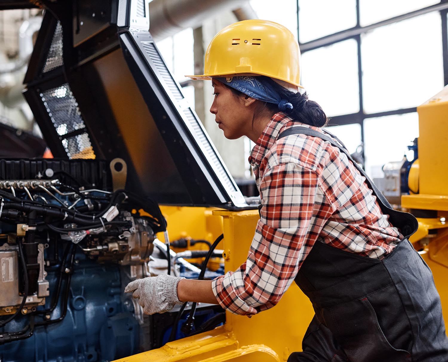 Young female technician in coveralls and hardhat checking motor of industrial machine while working in large modern factory